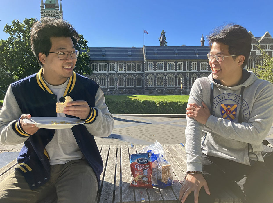 two men sitting on a bench in front of the clocktower, one of them holding a sandwhich