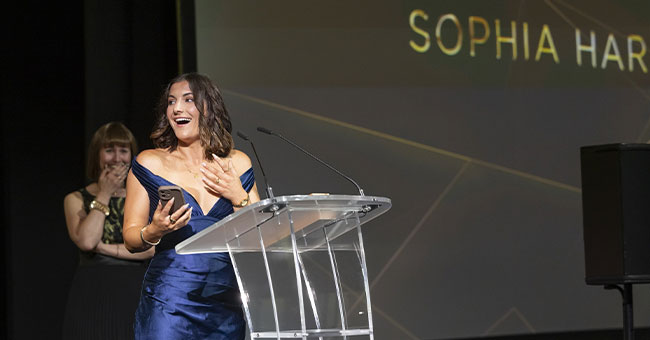 A woman in a blue dress standing at a podium with another woman in a black dress looks on.