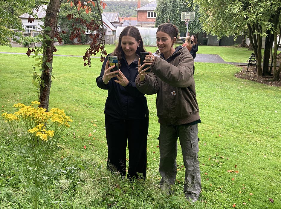 Two young women take photos of plant life with a mobile phone.