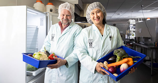 two people standing in lab coats, next to an open fridge and holding containers with vegetables in them