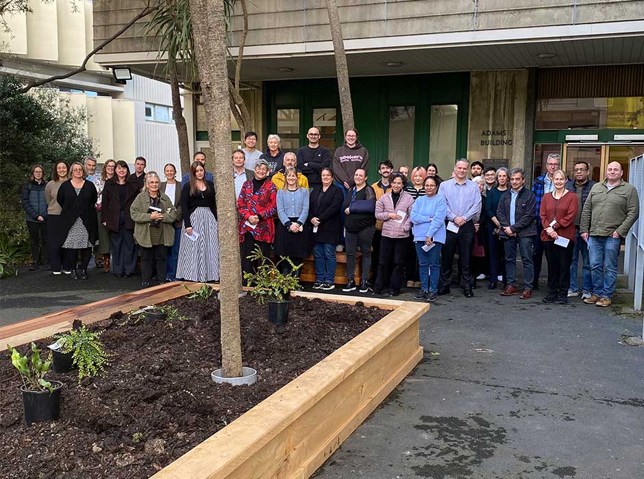 a row of people standing in front of a concrete building but behind a newly made raised garden bed with a few plants in it