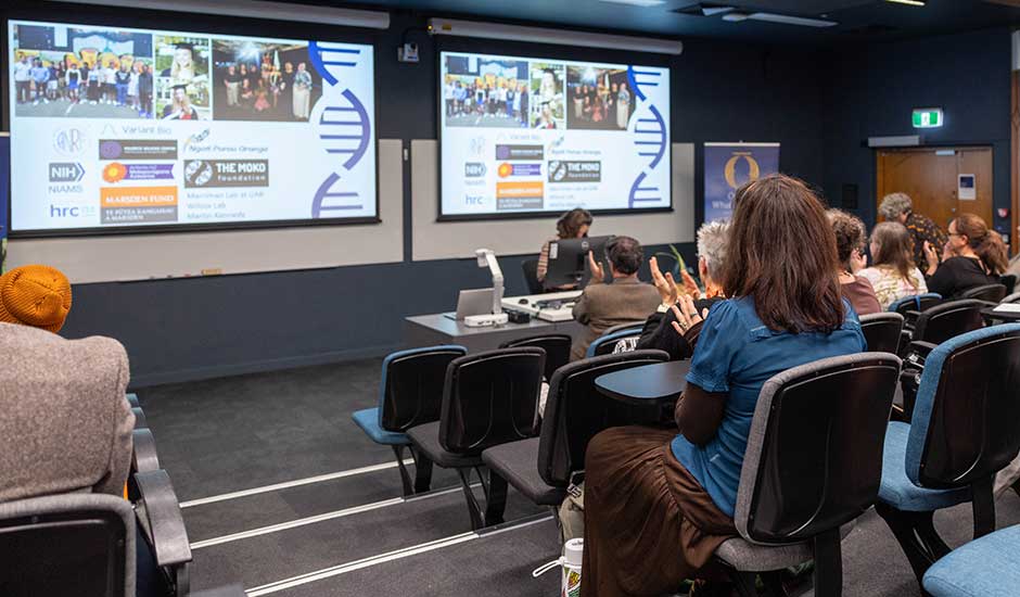 people seated in a lecture theatre, a person at the front presenting with two screens up behind them