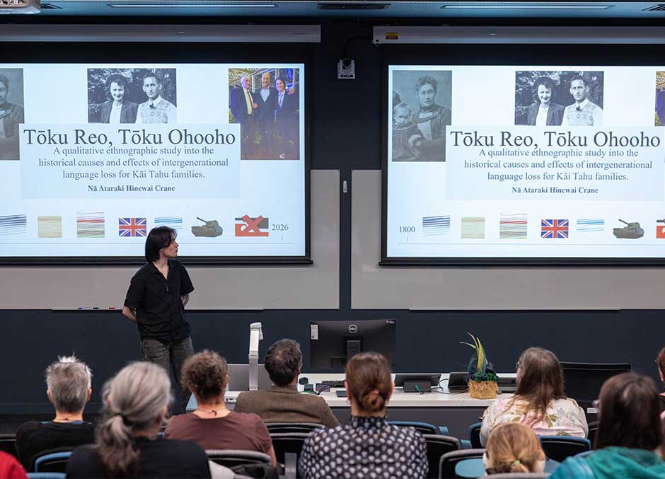 people seated in a lecture theatre, a person at the front presenting with two screens up behind them