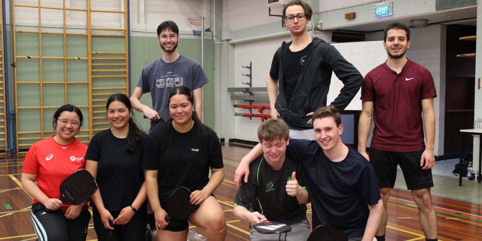 Eight casually dressed male and female students with pickleball bats posing in a gymnasium.