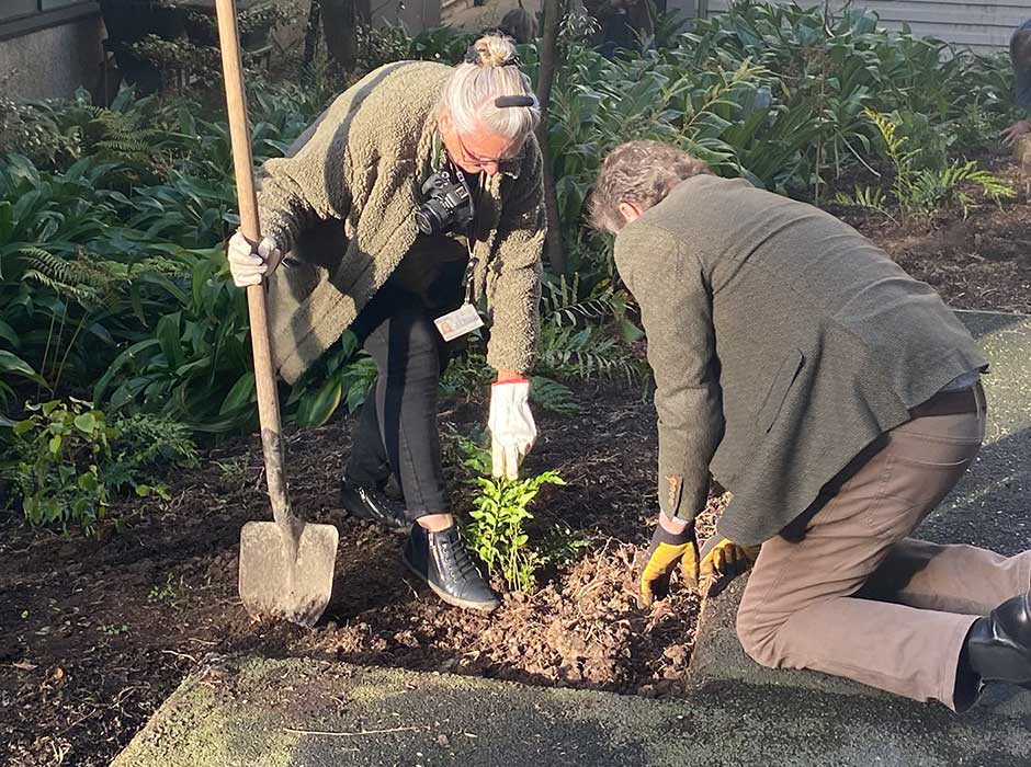 a person bent down with a shovel, and another person kneeling, while putting a plant in the ground