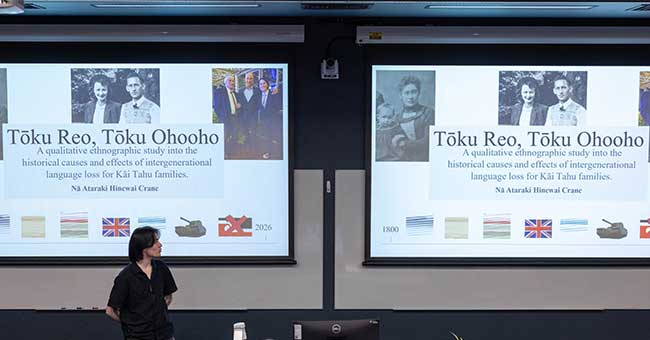 people seated in a lecture theatre, a person at the front presenting with two screens up behind them