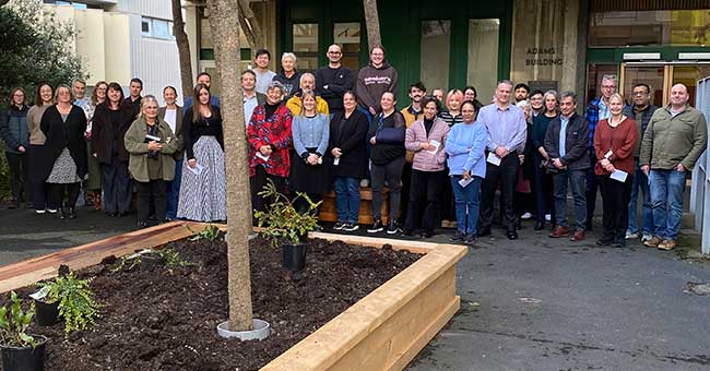 a row of people standing in front of a concrete building but behind a newly made raised garden bed with a few plants in it