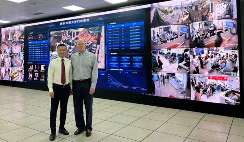 Two men stand inside a building, there is large illuminated screen behind them with photos and information on it