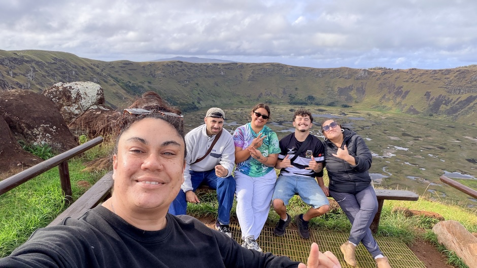 Local community members and University of Otago delegates pose for a selfie at a lookout on Rapa Nui. In the foreground, one participant holds the camera, while four others sit on a viewing platform behind. The background shows a large volcanic crater with steep grassy slopes, wetlands across the crater floor, and a cloudy sky overhead.