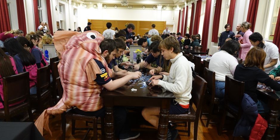 A large room full of students in groups at tables, putting together jigsaw puzzles – one is dressed as a fish