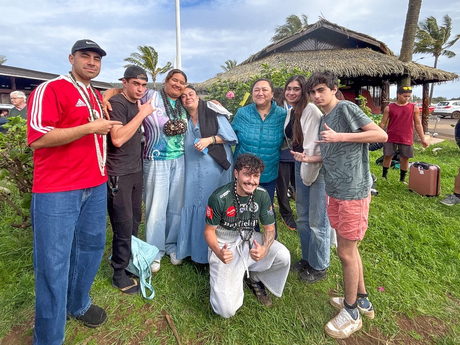 Group photograph taken outdoors on Rapa Nui. Several members of the University of Otago delegation stand alongside local community members on a grassy area in front of a traditional thatched-roof building. Palm trees bend in the wind in the background, and some participants wear flower garlands or necklaces. One person kneels at the front of the group while others stand close together for the photo.