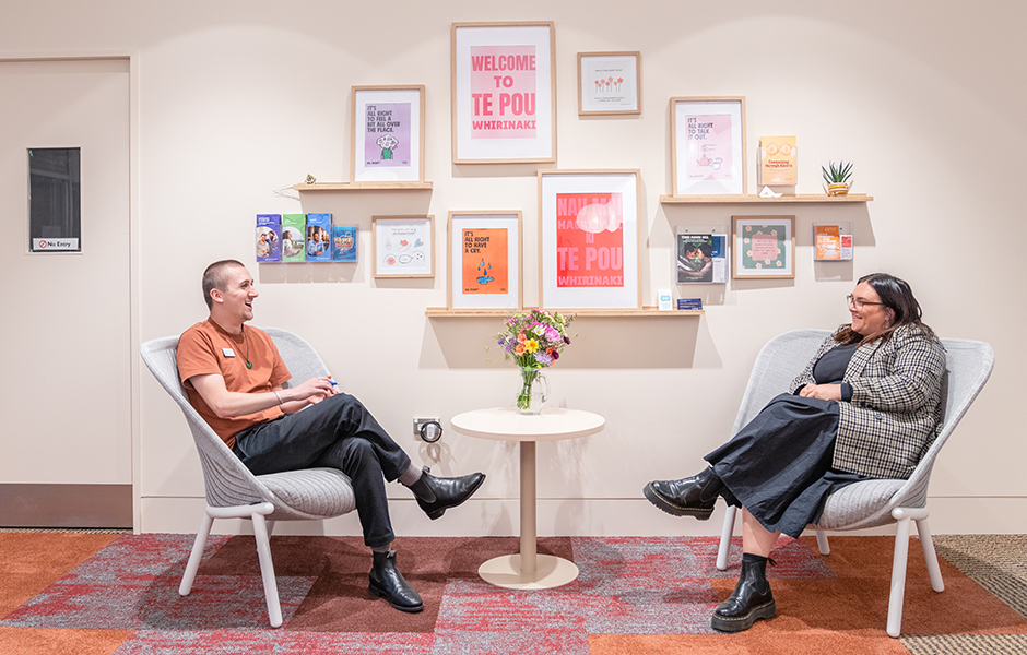 Two people sitting at a table in front of wall hung picture frames at the Student Wellbeing Hub.