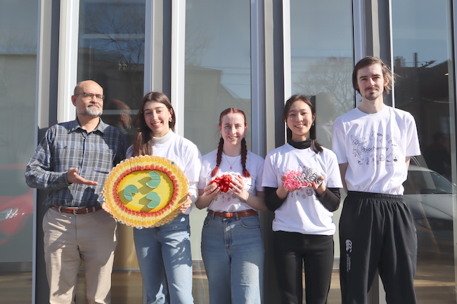 Four students wearing biochemistry branded t-shirts and their professor stand in front of large windows, two holding plastic protein models, and one holding a cardboard representation of a lysosome.