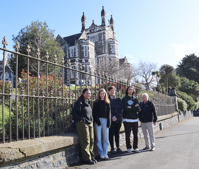 Four students and their professor stand lined up in front of an old iron fence and a castle-like stone school building on a blue-sky clear winter day