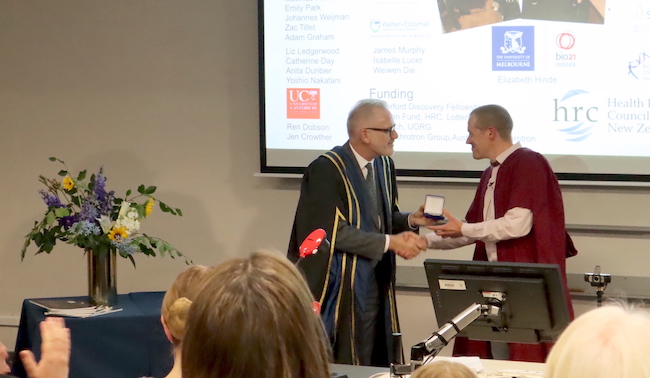 Two men in academic regalia standing in front of a seated audience, one handing a small box to the other while smiling and shaking hands.