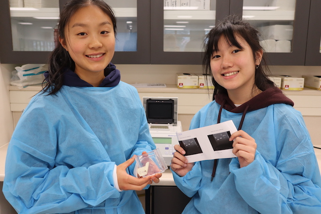Two smiling female students dressed in blue lab coats standing in a teaching lab, one holding an agarose gel, the other holding a photo of the DNA bands from the gel.