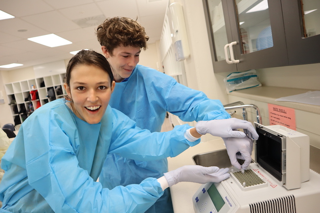 Two smiling students with blue lab coats on put microtubes into a PCR machine.