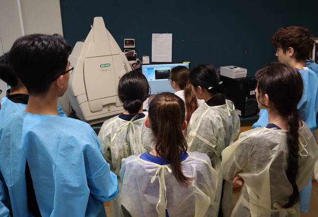 A group of students in blue or yellow lab coats look away from the camera towards a screen showing a photo of a DNA gel.