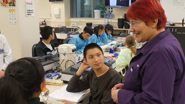 A biochemistry teaching fellow with red hair and a purple lab coat on talks to students sitting down at a work bench in a teaching lab