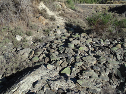 An erosional gully on the slopes of Tucker Hill, that has been exposed by historic mining. Schist bedrock in the floor of the gully (exposed in left foreground) is now covered with mined remnants of rounded boulders of greywacke from an old uplifted Manuherikia River channel. Some angular slabby schist boulders are also present, attesting to minor local bedrock erosion. A large brown silcrete boulder (left of centre in background) attests to erosion of gold-bearing 20 million year old quartz gravels on the slopes of Tucker Hill as well. Rail Trail passes in background.