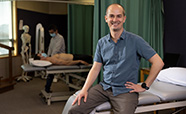 Daniel Ribeiro standing in a examination room with a patient lying on a bed in the background