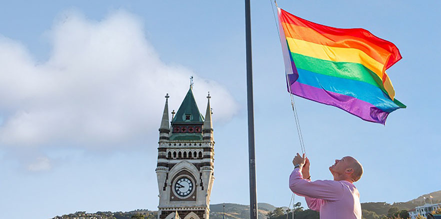 Man raising rainbow flag in front of Otago University clocktower